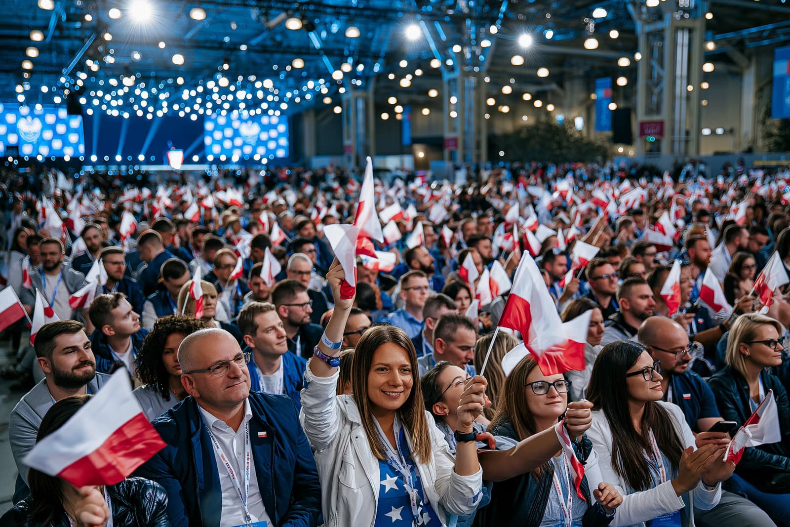 AfD'nin Magdeburg kampanya lansmanında, sert etkinlik ışıkları altında küçük bayraklar sallayan tıklım tıklım dolu bir sergi salonu ve uzakta görünen bir sahne.